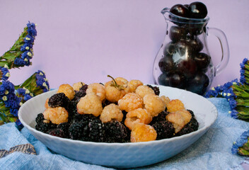 Frozen yellow raspberries and mulberries on a plate along with a jug of sweet cherries and flowers