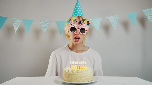 Happy smiling young girl in glasses with happy birthday lettering blows out candles on birthday cake