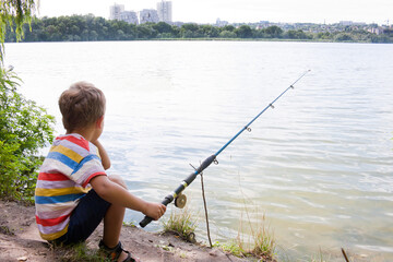 child is fishing in river on sunny summer day. young fisherman boy cast a fishing rod and is sitting waiting for a bite, view of river. Nature. Outdoor. Happy and interesting childhood