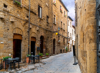 A picturesque narrow alley with sidewalk cafe tables in the historic medieval center of the Tuscan hill town of Volterra. - obrazy, fototapety, plakaty