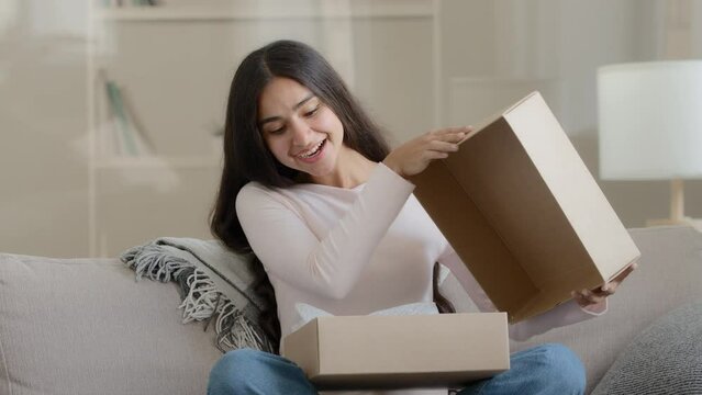 Surprised arab latin woman satisfied customer shopaholic sits on couch at home open delivery parcel postal carton box unpack present gift look at shipping goods purchases from internet clothing store