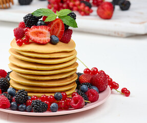 Stack of baked pancakes with fruits in a round plate on a white table