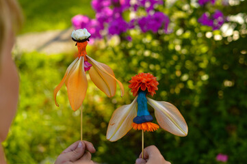 Close-up of children's hands holding dolls made of flower petals and plasticine on a stick. Ideas...