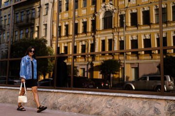Fototapeta na wymiar Side view of stylish woman a black dress and jeans jacket strolling along street near a vintage building . Modern lady in sunglasses walks in city . Street style shooting. Women's fashion . 
