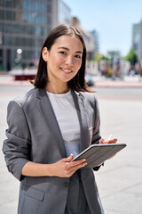 Young happy Asian business woman entrepreneur wearing suit holding digital tablet standing on big city busy street using smart business software tech for online work on pad computer outdoor, vertical.