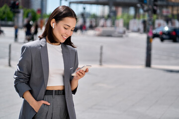 Young smiling elegant Asian busy business woman leader wearing suit standing in big city using cell phone platform applications. Smiling woman holding smartphone walking on street outdoors. 