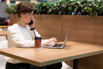 young woman sitting at the table talking on the phone typing something in the laptop
