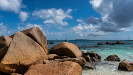 Fototapeta na wymiar Huge picturesque boulders on the coast of the Indian Ocean are washed by turquoise water. Ships on the horizon. Clouds in the blue sky. Seychelles. Praslin Island. Anse Lazio beach