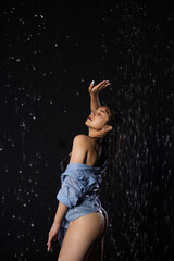 Beautiful girl in a black swimsuit and shirt poses in the rain, enjoying the drops of water. She stands with her hand raised, her eyes closed. Water splash on black background. Aqua studio