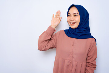 Smiling young Asian muslim woman in pink shirt gesturing wave hand isolated over white background. People religious lifestyle concept