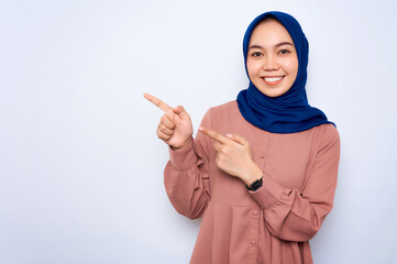 Smiling young Asian muslim woman in pink shirt pointing fingers at copy space isolated over white background. People religious lifestyle concept