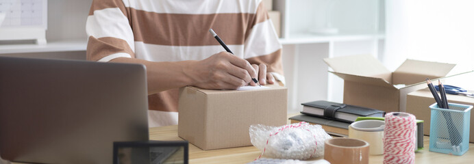 Young Asian man is writing down the customer's details and addresses on the notebook or box in order to prepare for shipping according to the information, packing box, Sell online, freelance working.