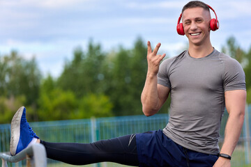 Joyful athlete smiles while stretching his legs at stadium, listening music with headphones. Runner...