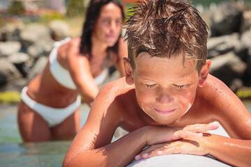 Cheerful teenage boy lying on a swimming board. Happy european family having fun floating on a swim...