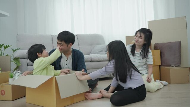 Parents and children play with brown cardboard boxes as they move into their new home. Asian family smiling and laughing happily in house. relocation,  move in-out, buying, new furniture, moving day.