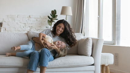 Cheerful happy mom cuddling cute little daughter kid on sofa, tickling, caressing laughing...