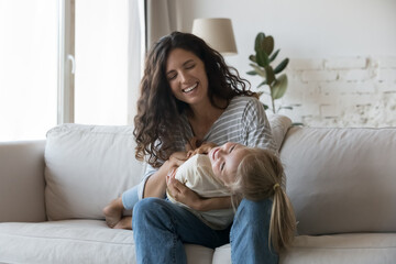 Happy joyful mom tickling cheerful cute little daughter, holding laughing kid in arms in lap,...
