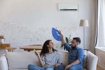 Young couple frustrated with heat, waving handheld fan, using remote control, turning air conditioner on for cooling air, sitting on couch at home. Domestic equipment failure concept © fizkes