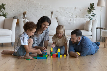 Happy parents and little adopted children playing in heating floor together, building towers from...