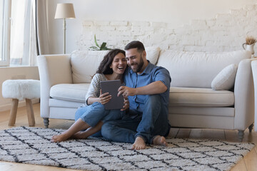 Happy laughing married couple using tablet, sharing digital device on heating floor at home, browsing internet, shopping online, using ecommerce app, making video call, enjoying leisure, communication