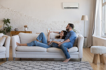 Happy millennial couple of homeowners enjoying cool conditioned air, resting on couch together,...