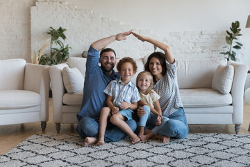 Happy millennial family couple and two little children hugging under hand home roof, sitting on heating floor in cozy living room, smiling at camera. Real estate, house buying, insurance concept