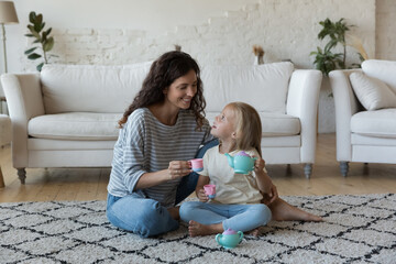 Happy playful caring mother and sweet little daughter girl playing tea party with tiny toy cups, teapot on heating floor, pretending drinking coffee in game at home, talking, laughing