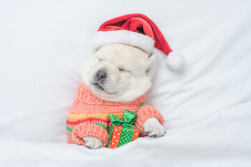 Funny Golden retriever puppy wearing warm sweater and red santa's hat sleeps under white blanket on a bed at home and hugs gift box. Top down view