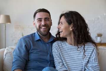 Cheerful millennial couple home head shot portrait. Happy husband and wife sitting on couch, looking at camera, laughing, talking on video call, chatting. Communication, relationship, love concept