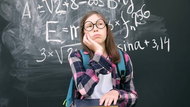 Teen girl little student in glasses and blue backpack standing and thinking on chalkboard background. Then she is pointing up with finger because finds answer. Brainstorming and Bright idea concept.