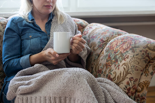Close up of hands holding blue cup of tea or coffee. Model and cozy plaid is sitting, relax at home on sofa