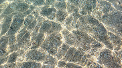 Sea sandy bottom is visible through clear turquoise sea water. Sun glare on sea surface. Beautiful abstract background. Copy space. Selective focus