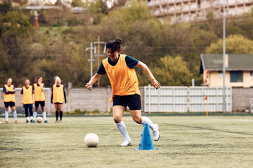 Female soccer player dribbling ball around cones during sports training.