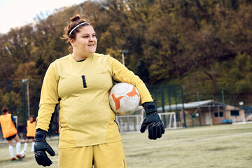 Female goalkeeper holds ball while standing on soccer field and looking away.