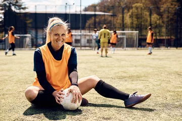 Gordijnen Happy female soccer player relaxes on grass on playing field and looking at camera. © Drazen