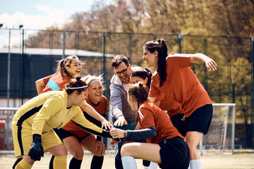 Cheerful female soccer players and their coach gathering hands in unity before the match on playing field.
