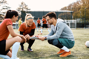 Female players and their coach going through game strategy before soccer match on football field.