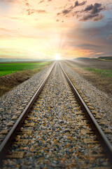 Vías del tren a través de campos de cultivo, que convergen hacia el sol en el horizonte - obrazy, fototapety, plakaty