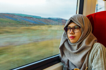Portrait of young and beautiful Asian Muslim woman wearing eyeglasses and hijab sitting alone against the window in a moving train. Looking out through window. Contemplating expression.
