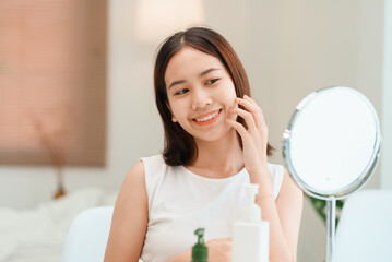Closeup portrait of smiling young beautiful asian woman looking at mirror touching her skin enjoying treatment for dry skin. 