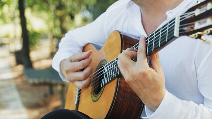 Professional guitarist plays guitar outdoors. Musician plays a classical guitar in the park. A man in a white shirt plays a musical instrument outside the house. The guitarist plays the guitar. - obrazy, fototapety, plakaty