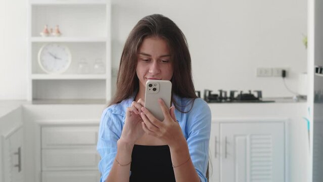Happy attractive girl looking at smartphone relaxing on chear at home, enjoying using online mobile apps technology, playing games on cell phone, checking messages or social media posts