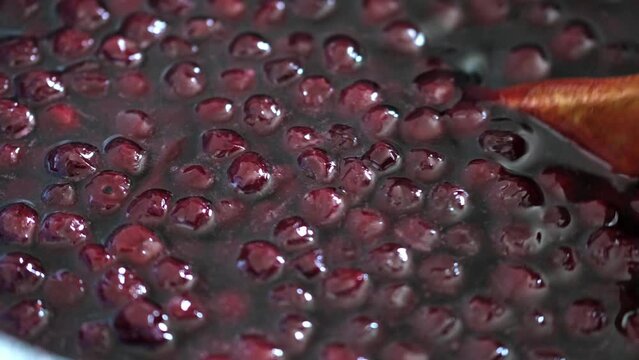 Cooking pot with sweet cherry jam close-up. Stirring boiling confiture with wooden spoon