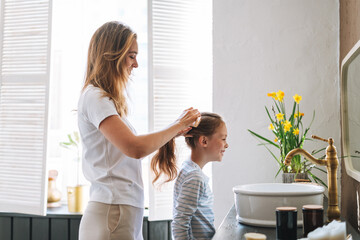 Young mother woman with long hair with little tween girl daughter in pajamas combs her hair on...