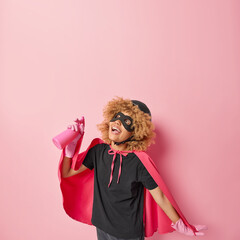 Positive busy female cleaner dressed in superhero costume holds detergent focused above does spring cleaning going to dust ceiling isolated over pink background blank space for your promotion