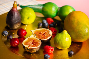 A golden plate, platter of fresh tropical fruit stands on a table. Figs cut in half, mango, limes and seasonal blue berries, raspberries. Variety of summer juicy fruits still life close up. Warm light