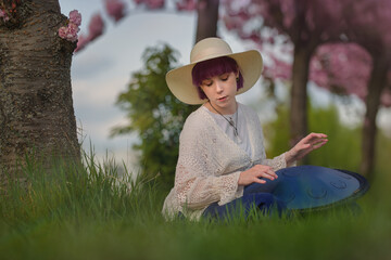 A young purple-haired woman sitting in a flowering orchard plays a tongue drum. It is a musical instrument similar to handpan, hang or pantam.