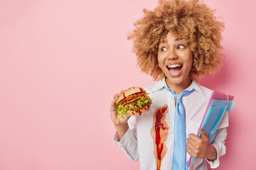 Positive woman with curly hair dressed in white formal shirt and tie eats appetizing burger holds folders with sticker looks gladfully away isolated over pink background empty space for your promo