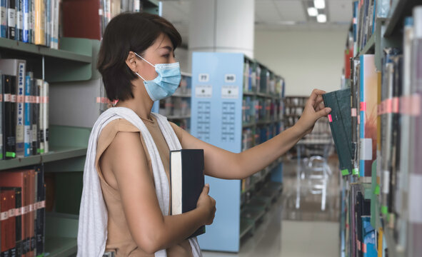 Woman with face mask searching data and select book for research on shelf