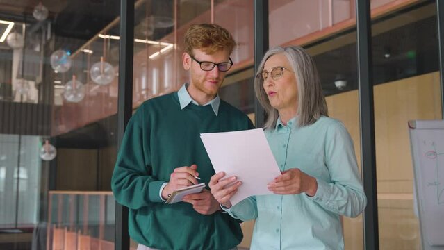 Mature businesswoman teaching on documents young male intern in office